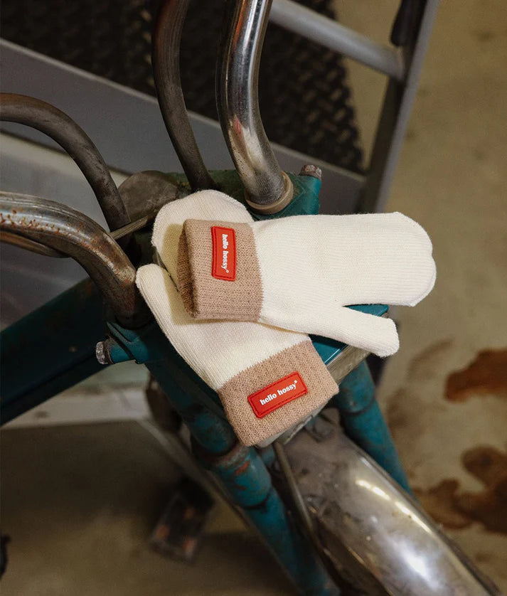 Par de luvas Hello Hossy Hotty Sandy creme com punho castanho e etiqueta vermelha, pousadas no guiador de uma bicicleta.