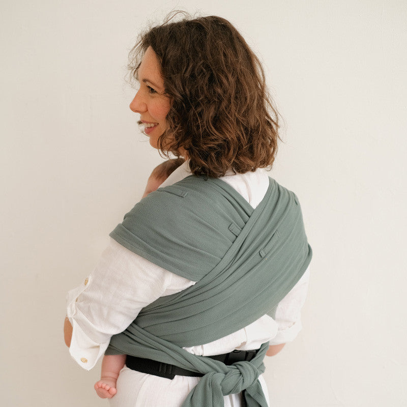 Mãe a ajustar o Mei Tai Evolutivo Neobulle P'tit Evo Eucalyptus nas costas, com o bebé virado para si. A mãe está de perfil e o pano é de cor verde.