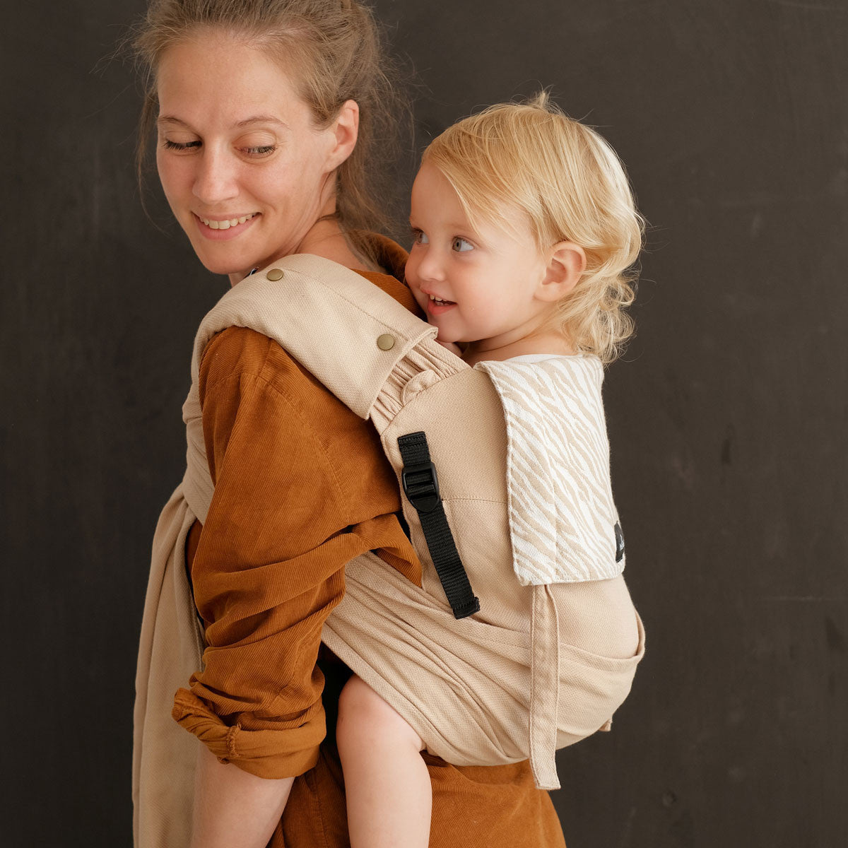 Uma mãe a carregar o seu bebé nas costas, demonstrando a versatilidade do Mei Tai Evolutivo Neobulle P'tit Evo Savane.