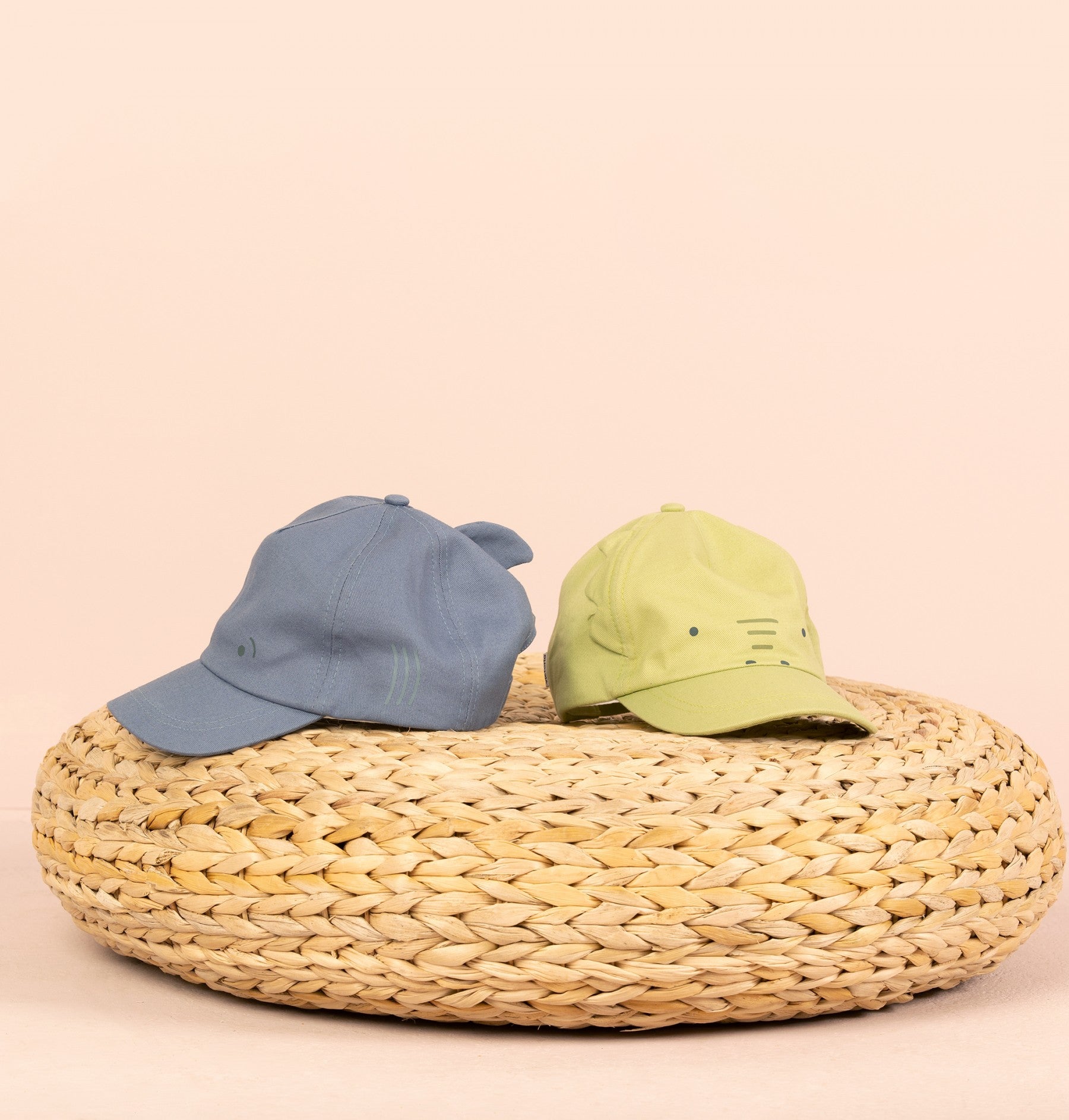 Par de bonés infantis Victoria em lona, azul com tubarão e verde com crocodilo, em cima de um pufe de verga.