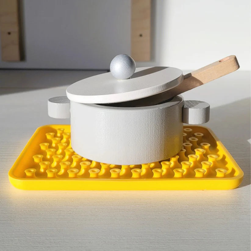 Uma base amarela do Wiggly Toy Jogo Sensorial a ser utilizada como salvamantéis, com um tacho de brincar cinzento e branco colocado em cima, demonstrando a sua versatilidade.