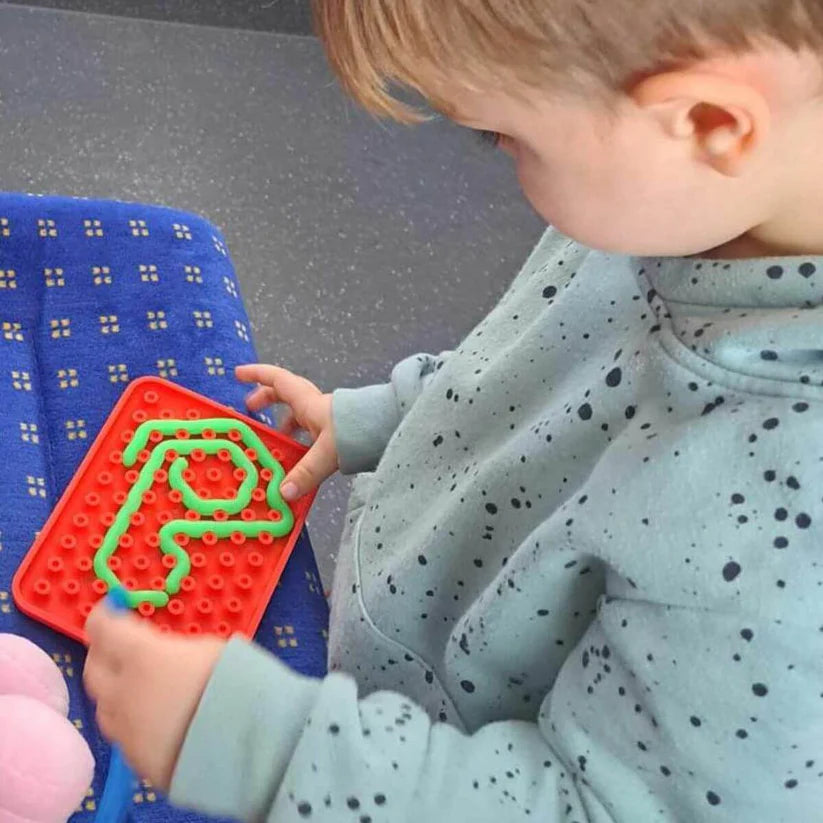 Uma criança pequena concentrada a brincar com um Wiggly Toy Jogo Sensorial vermelho, inserindo uma peça flexível verde na matriz para formar um padrão.