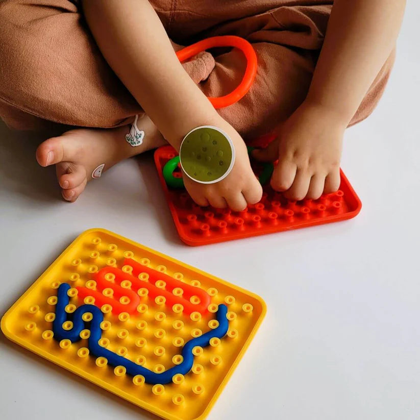 As mãos de uma criança a manipular as peças de um Wiggly Toy Jogo Sensorial vermelho, enquanto outro conjunto amarelo com peças azuis e cor-de-rosa está em primeiro plano.