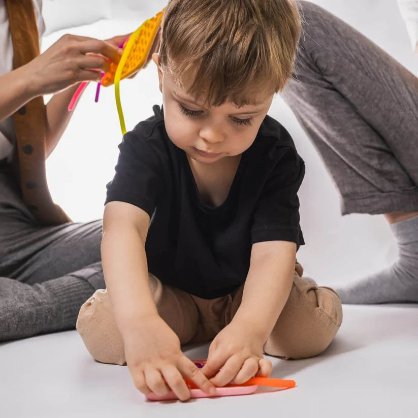 Um menino pequeno sentado no chão, focado em manipular e encaixar as peças do Wiggly Toy Jogo Sensorial, com adultos desfocados ao fundo.