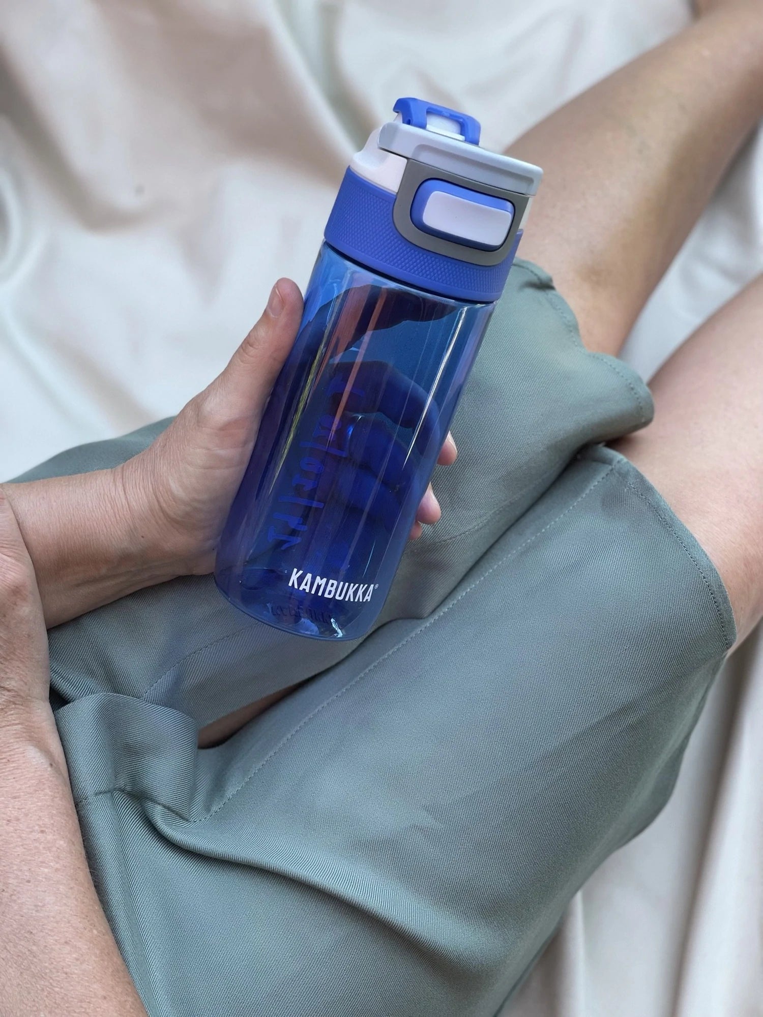 Mão a segurar a Garrafa Kambukka Elton 500ml azul transparente cheia de água, com as pernas de uma pessoa em fundo.