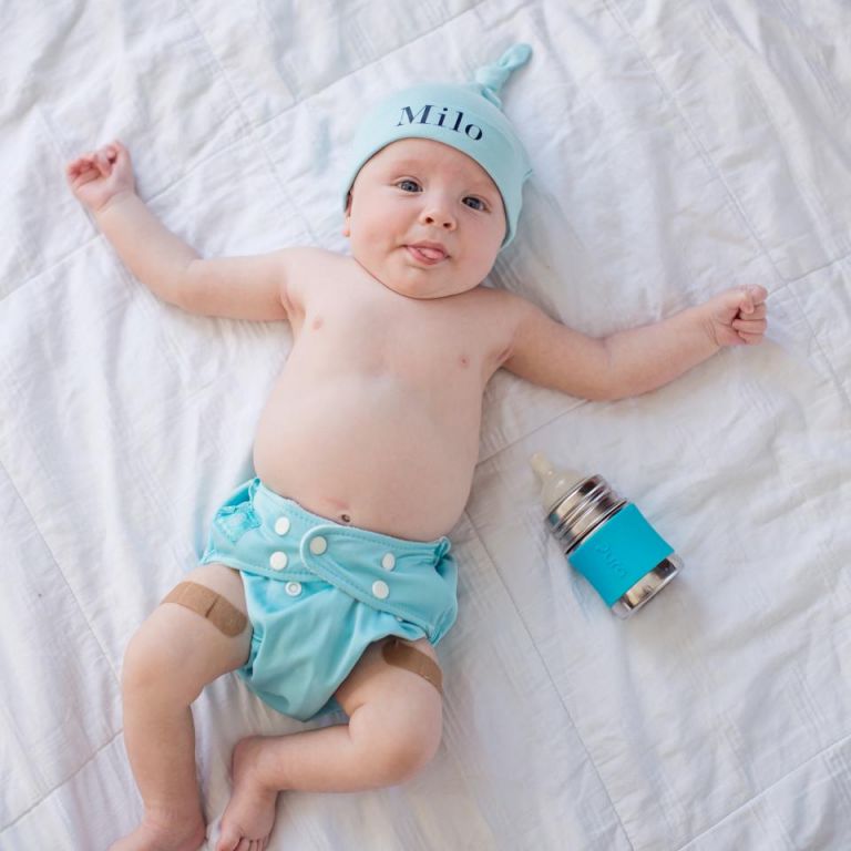 Bebé Milo deitado numa cama branca com fralda e gorro azul, e ao lado uma garrafa Pura de aço inoxidável com capa azul.
