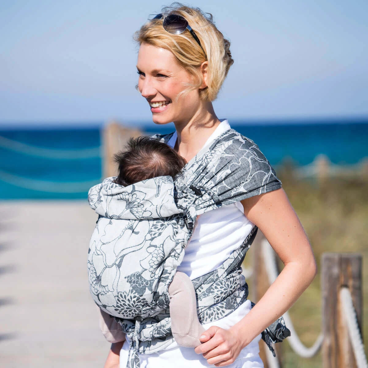 Mulher a usar o porta-bebé Mei Tai Hoppediz Evolutivo Hop Tye Buckle Florence, a caminhar na praia, com o bebé seguro e confortável no porta-bebé.