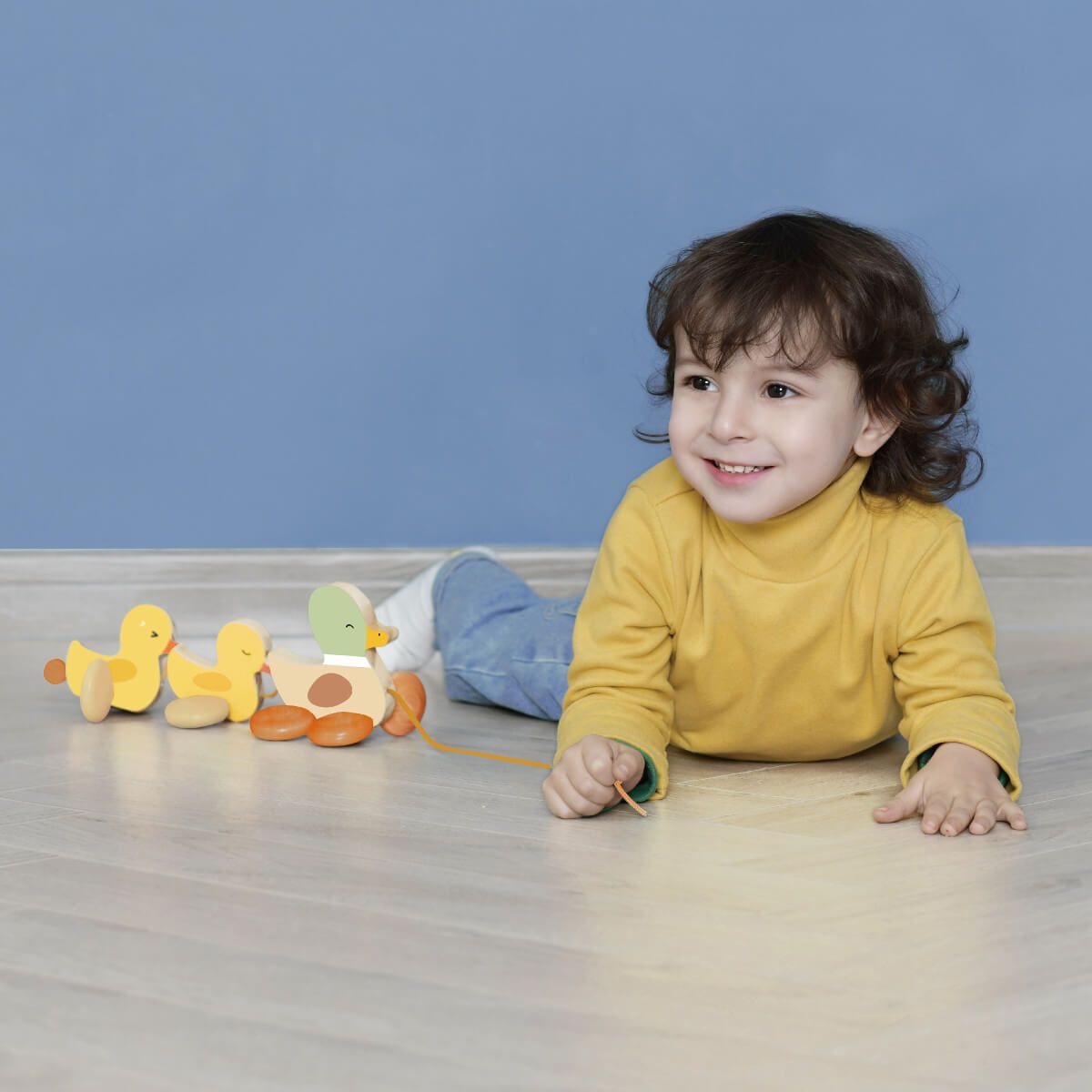 Menino sorrindo deitado no chão ao lado da família de patinhos de puxar Classic World, com a parede azul como pano de fundo.