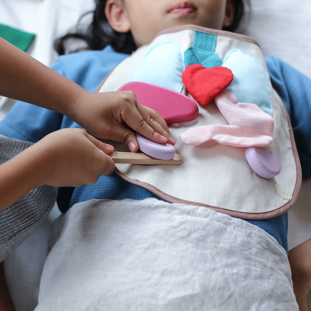 Close-up das mãos de uma criança usando o conjunto cirúrgico PlanToys, mostrando o detalhe das peças e a interação com o avental.