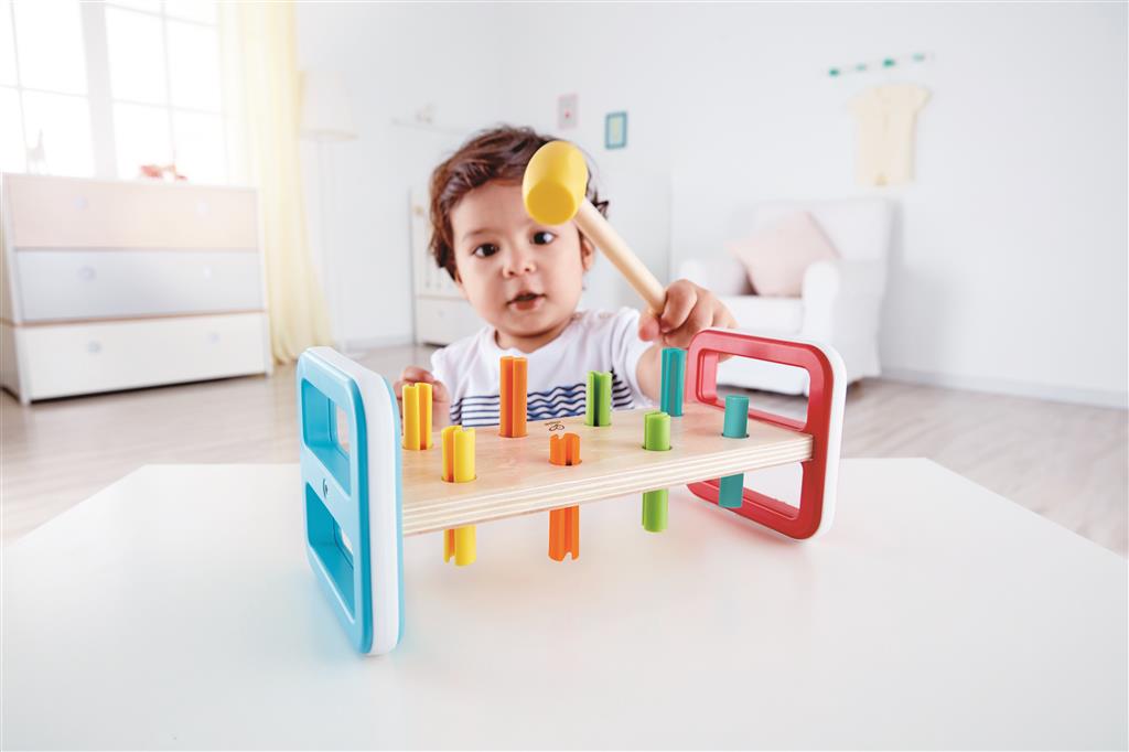 Bebé concentrado a brincar com o Jogo do Martelo da Hape numa mesa branca, segurando o martelo e pronto para bater num pino colorido.