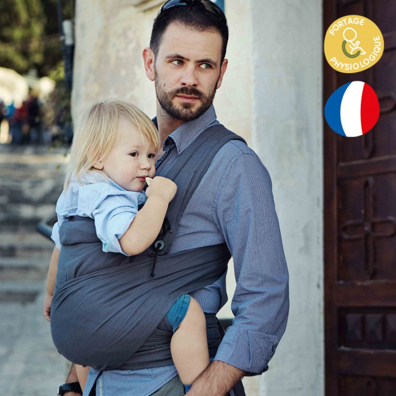 Pai de barba e camisa azul claro usando um mei tai cinza escuro, carregando um bebé loiro de cabelos compridos, parado em frente a uma porta de madeira antiga.