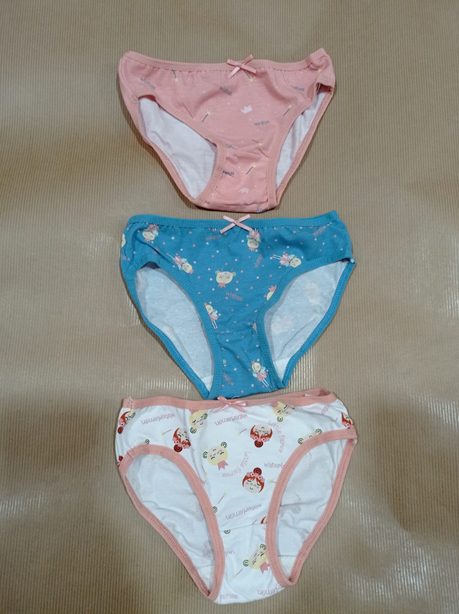 Três cuecas para criança, em tons rosa, azul e bege, sobrepostas numa superfície castanha. As cuecas mostram detalhes das estampas.
