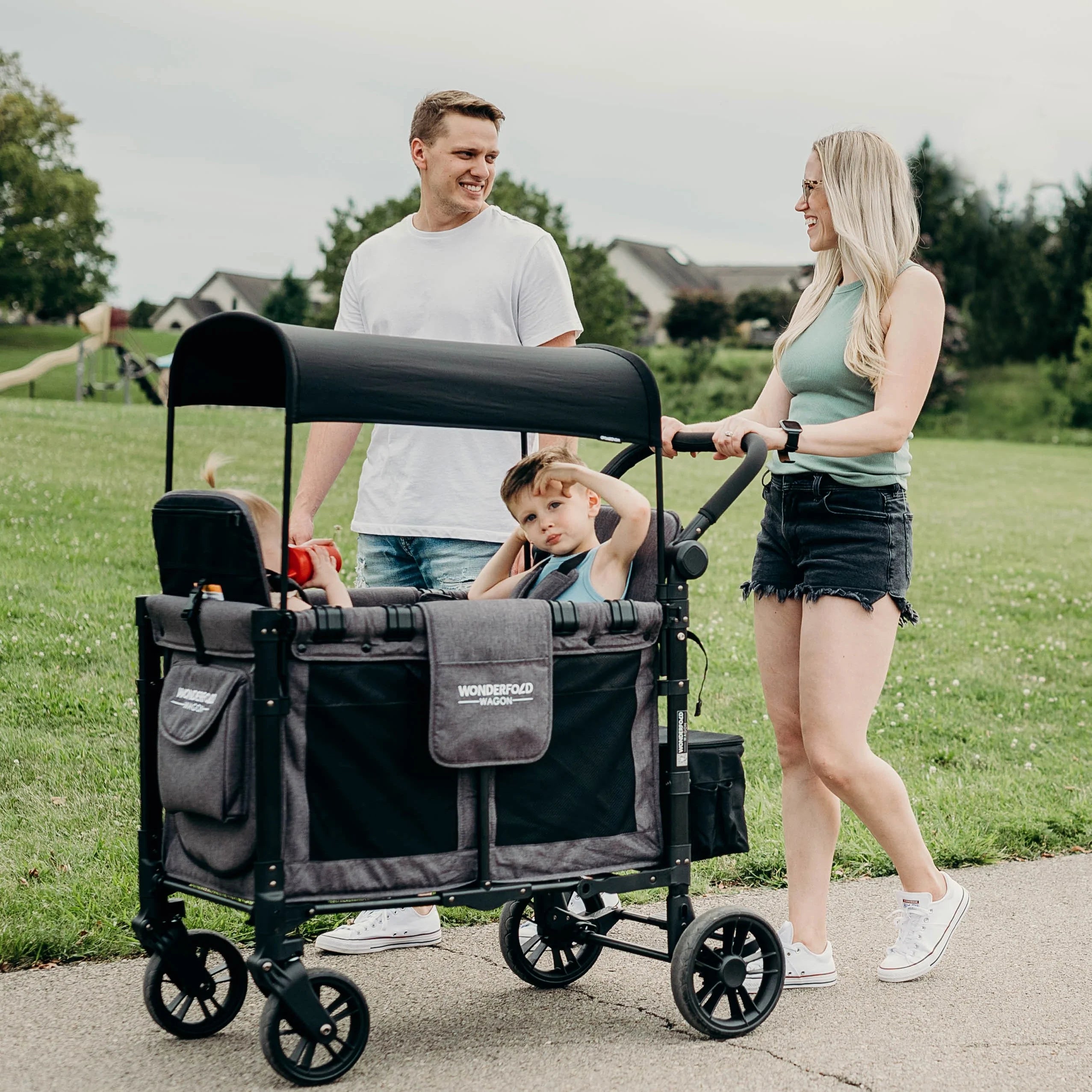 Família feliz usa Wonderfold Wagon Serie W para 2 crianças em passeio no parque. Carrinho vagão confortável e seguro.