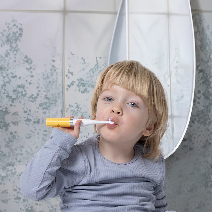 Criança pequena com cabelo loiro a escovar os dentes com a escova elétrica Matchstick Monkey Leão amarela, em frente a um espelho.
