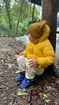 Fotografia de uma criança pequena sentada num ambiente de floresta, vestindo um casaco amarelo e usando botinhas barefoot combinando, explorando a natureza.