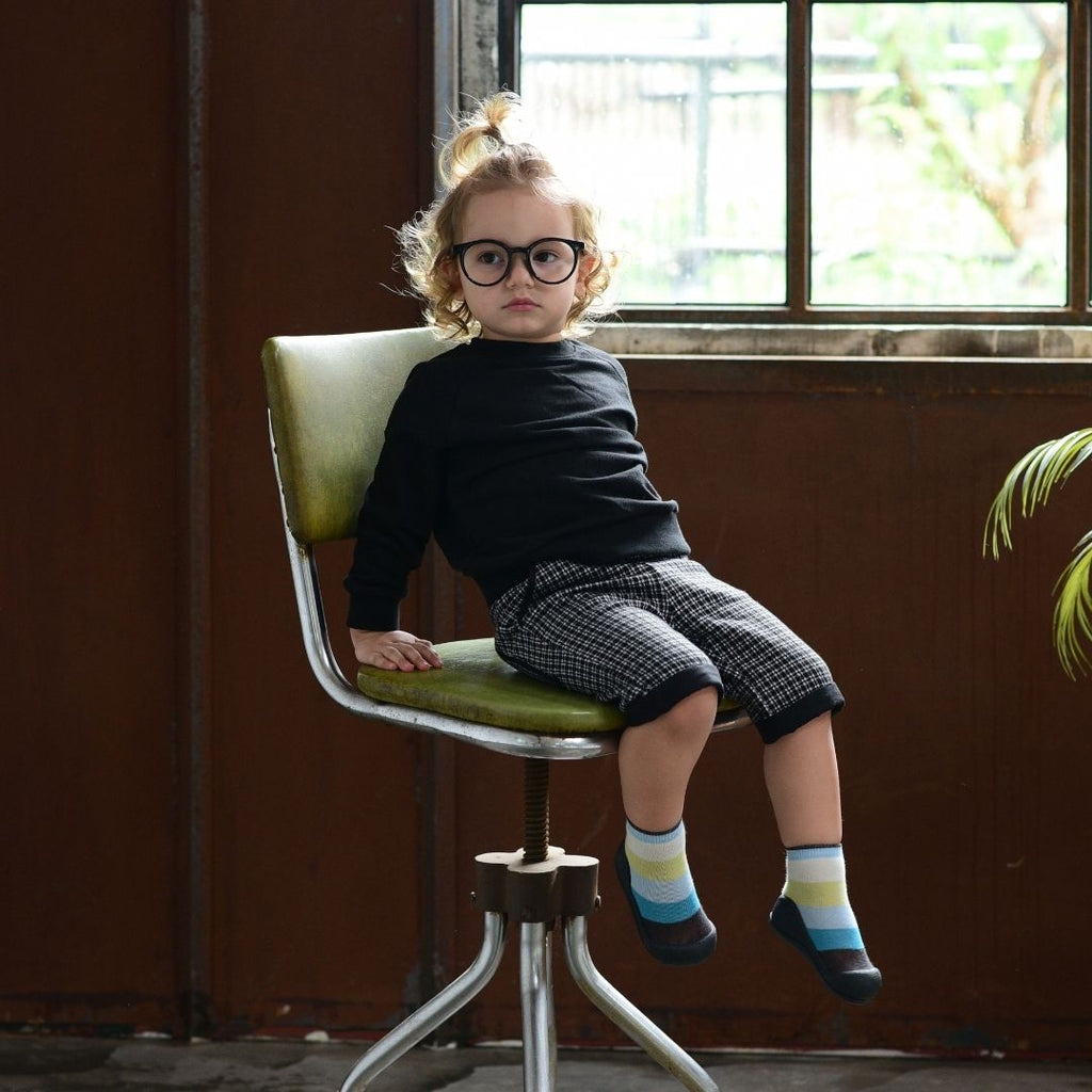 Criança sentada numa cadeira a usar os sapatos Attipas New Rainbow Blue, demonstrando o ajuste confortável e flexível do calçado.