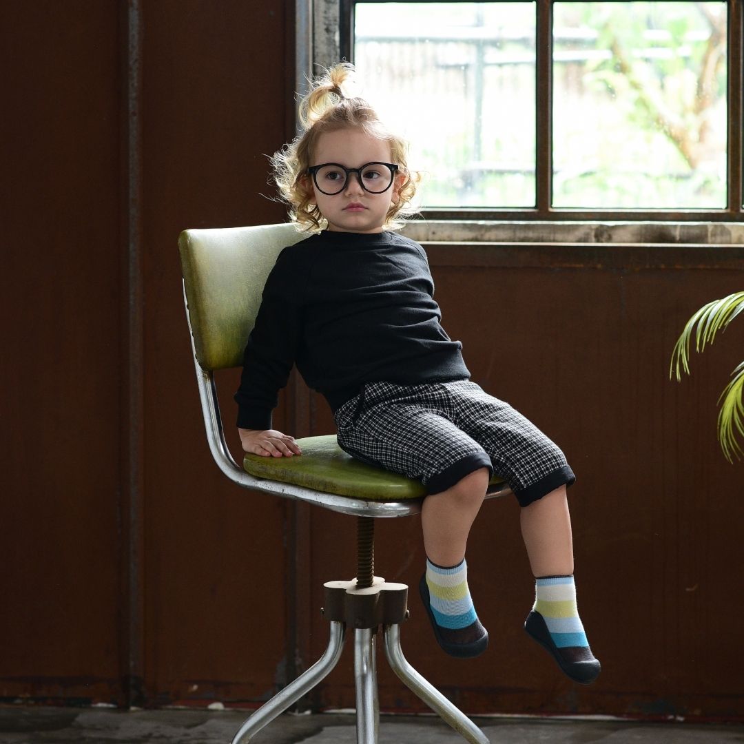 Criança sentada numa cadeira a usar os sapatos Attipas New Rainbow Blue, demonstrando o ajuste confortável e flexível do calçado.