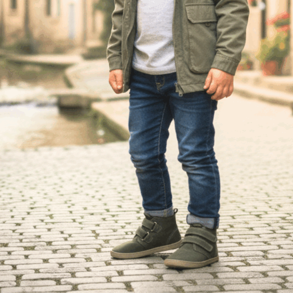 Criança com calças de ganga e casaco verde militar a usar as botas Baby Lobitos Barefoot Bota Militar em verde, de pé numa rua de calçada.
