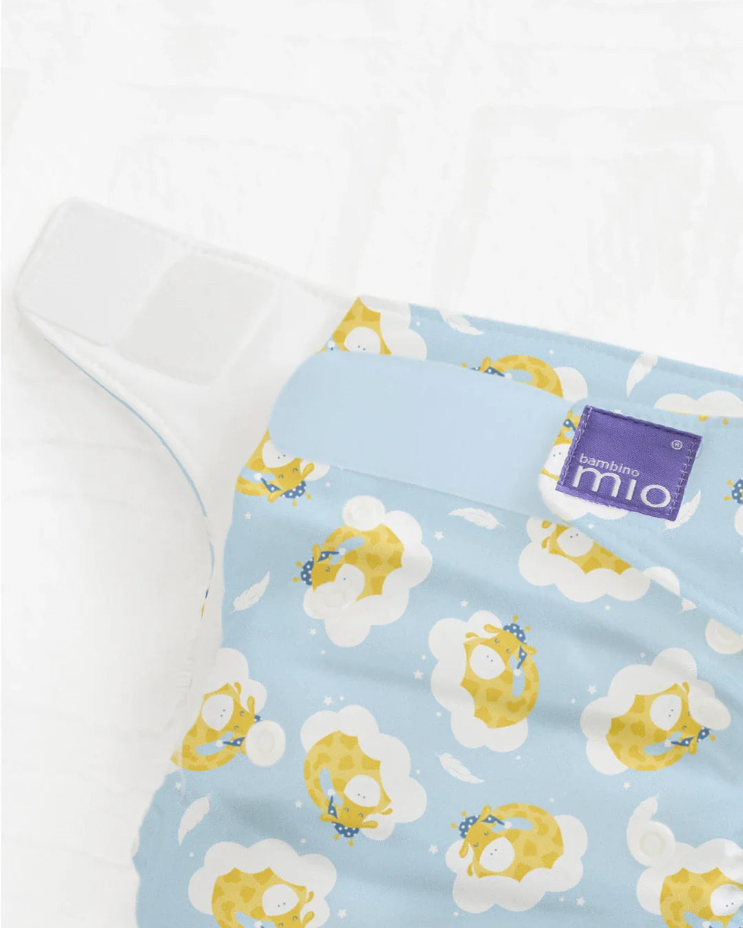 Detalhe de uma fralda reutilizável Bambino Mio miosolo classic com padrão de girafas em nuvens azuis, mostrando o fecho de velcro e a etiqueta da marca.