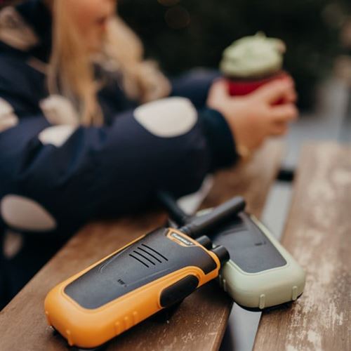 Dois walkie talkies KidyWolf, um laranja e um verde, sobre uma mesa de madeira, com uma criança desfocada ao fundo segurando uma chávena.