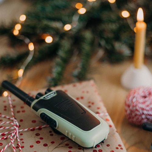 Um walkie talkie KidyWolf verde pousado sobre um presente embrulhado, com luzes de Natal e uma vela ao fundo, criando um ambiente festivo.