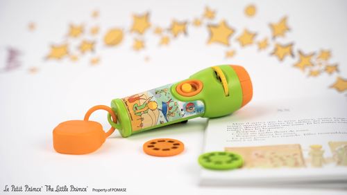 Lanterna projetor 'O Principezinho' pousada sobre uma página de livro, com um disco de projeção ao lado, num cenário claro.