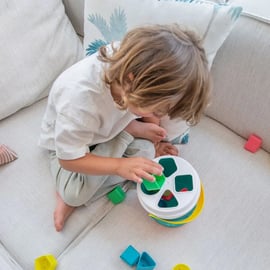 Criança a brincar no sofá com o Miniland Cubo com formas de encaixe, concentrada a encaixar uma peça verde.