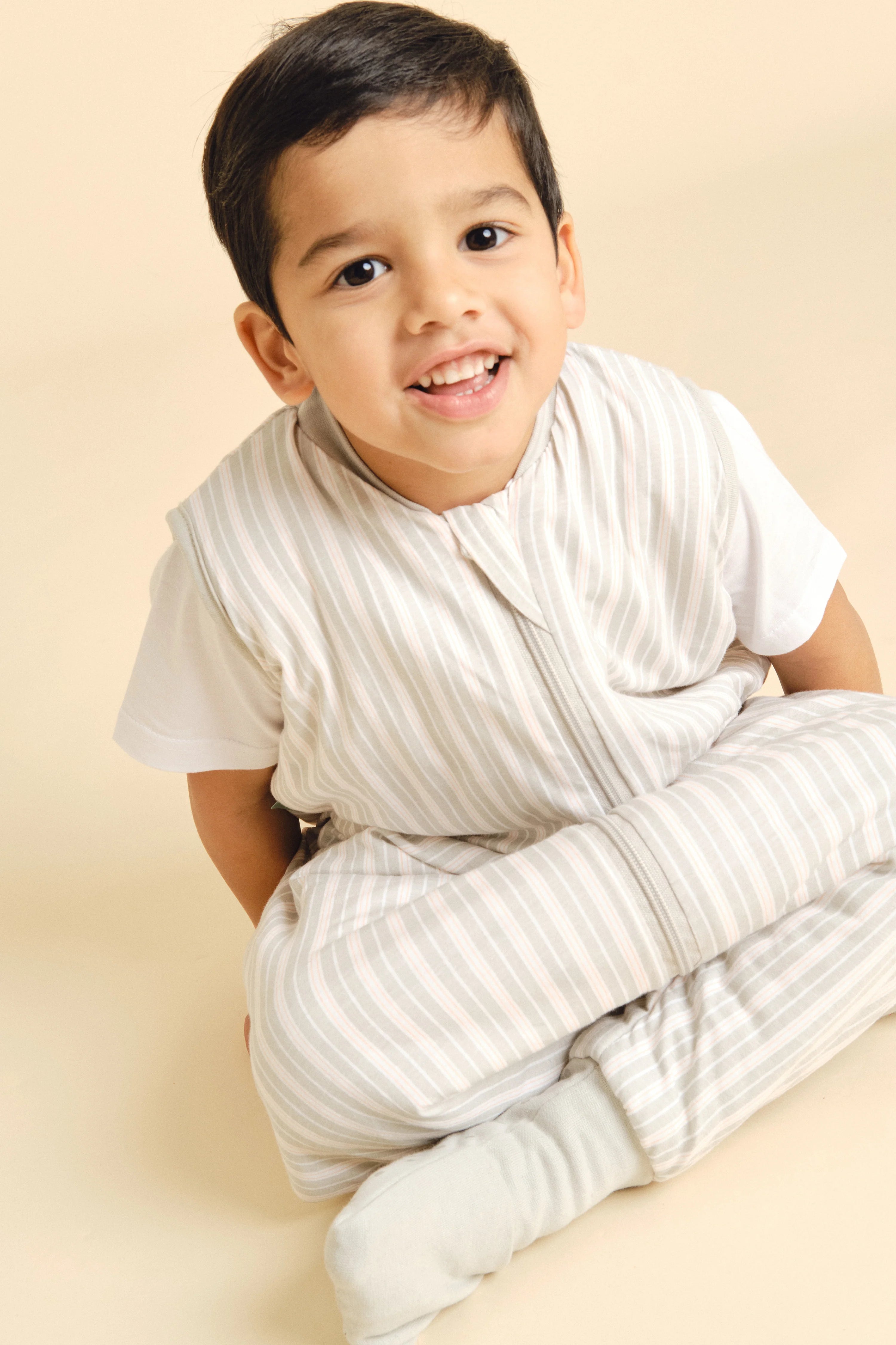 Criança sorridente com cabelo escuro, sentada, a usar o saco de dormir MolisandCo TOG 1.0 Dawn Stripes com pés e meias, que permite mobilidade.