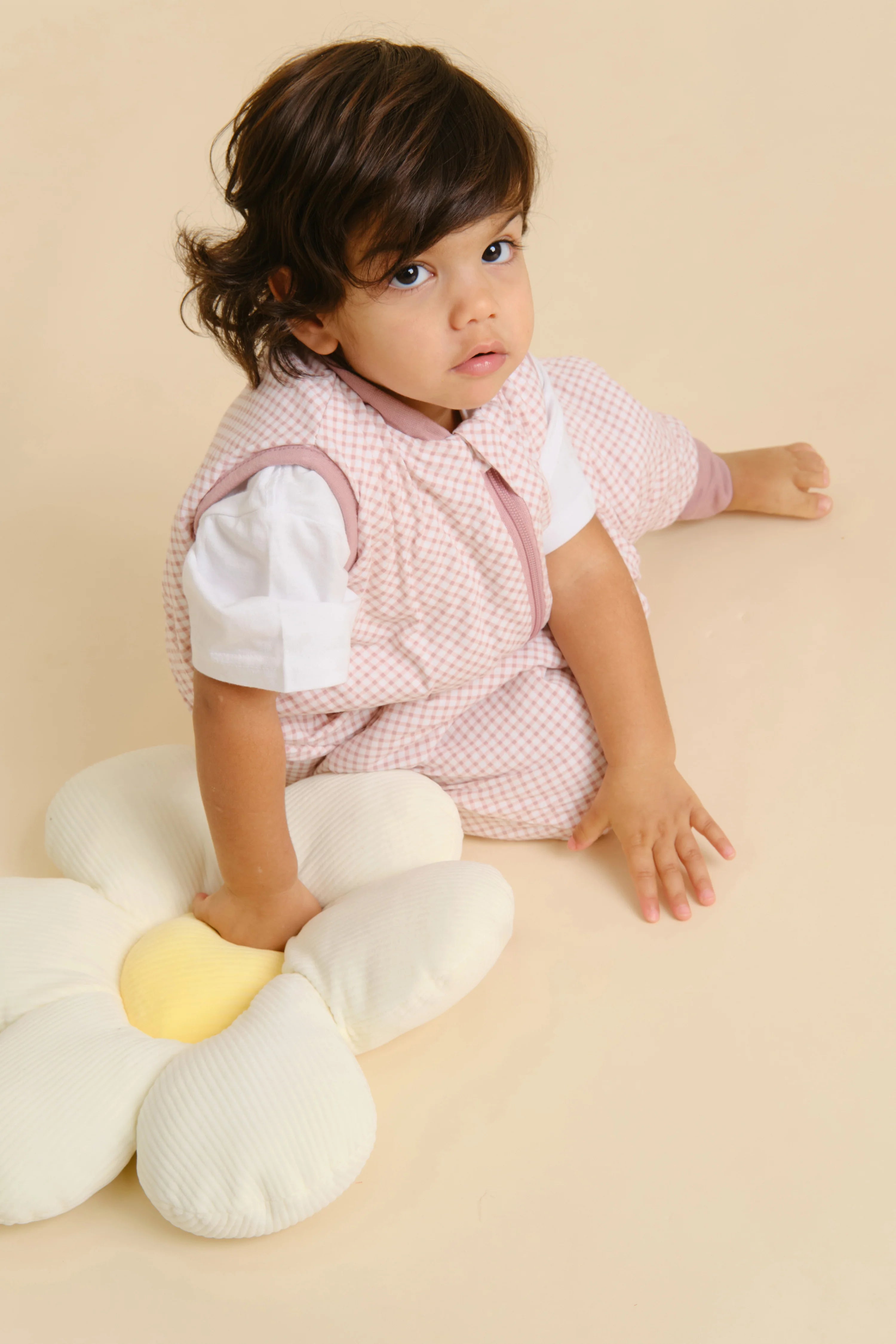 Bebé com o saco de dormir MolisandCo TOG 1.0 VICHY PINK, sentado no chão ao lado de uma almofada em forma de flor, olhando para a câmara.