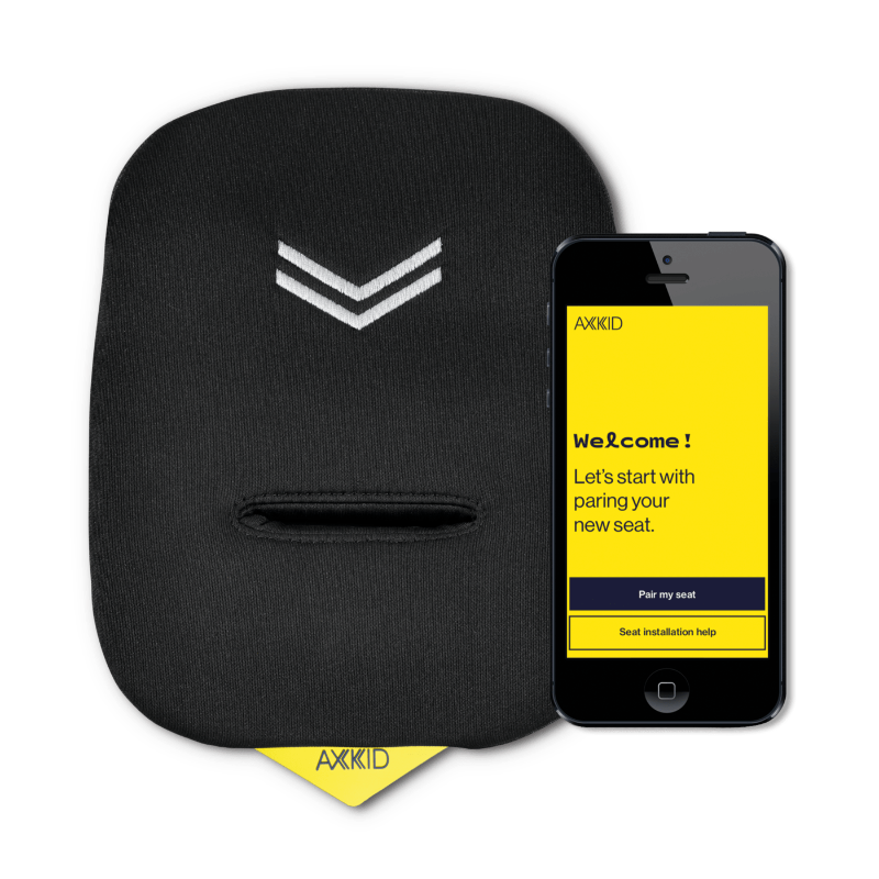 Almofada de segurança Axkid Connect ao lado de um smartphone que exibe a aplicação Axkid Connect. A almofada é preta com um pequeno detalhe em cinza e inclui uma abertura para a fivela de segurança. O smartphone mostra o ecrã inicial da aplicação em fundo amarelo.