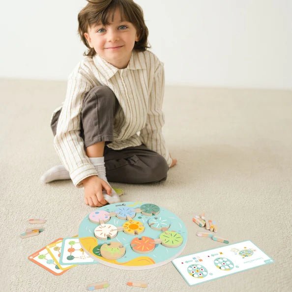 Menino sorridente a brincar no chão com o Classic World Jogo Flor de Lótus, demonstrando a interação com o tabuleiro e as peças de madeira.