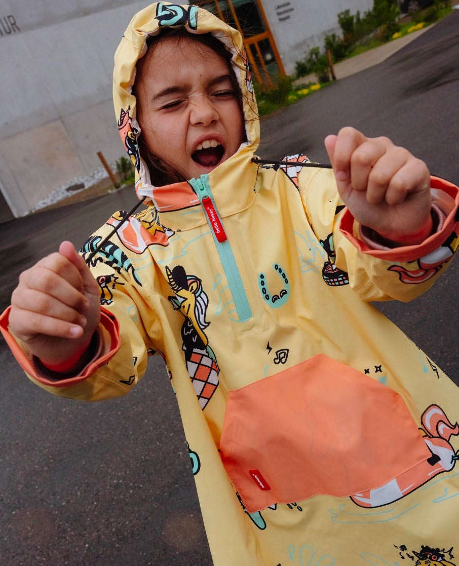 Menina com expressão divertida a usar o poncho Hello Hossy Unicorn amarelo com capuz, mostrando o padrão de unicórnios e o bolso laranja.