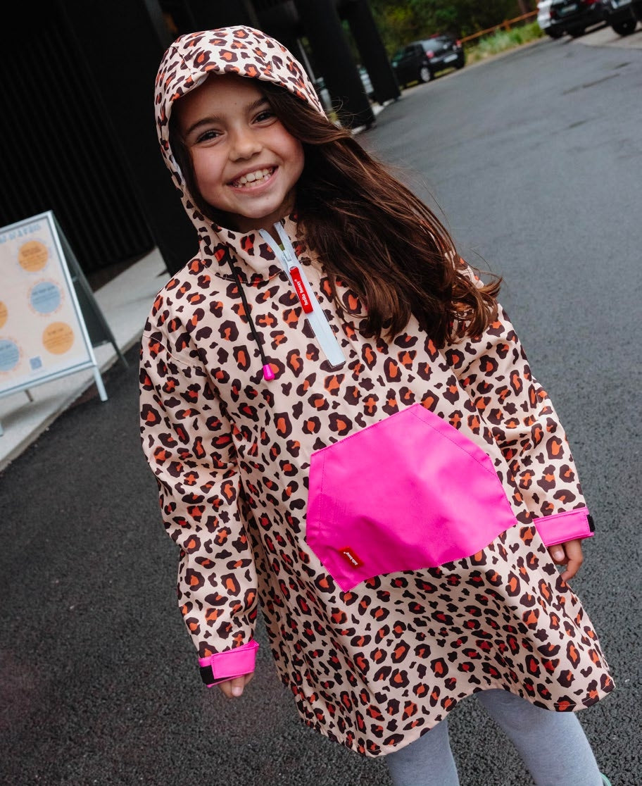 Menina sorridente a usar um poncho Hello Hossy Leopard #6 com padrão leopardo e detalhes em rosa néon, com capuz e bolso frontal.