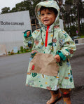 Criança sorridente a usar um poncho Hello Hossy Mountain Ride verde com padrões de montanha e veículos, capuz e bolso canguru, em dia de chuva.