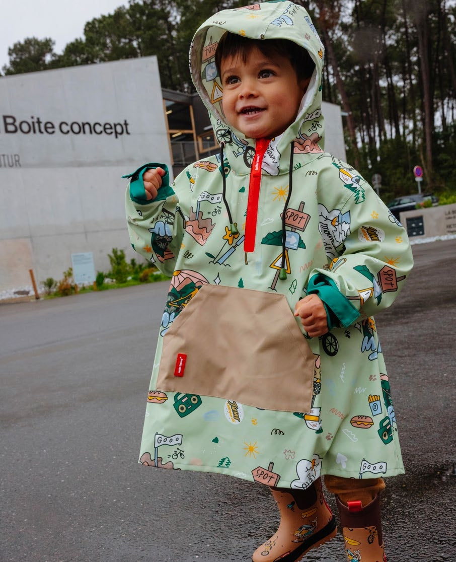 Criança sorridente a usar um poncho Hello Hossy Mountain Ride verde com padrões de montanha e veículos, capuz e bolso canguru, em dia de chuva.