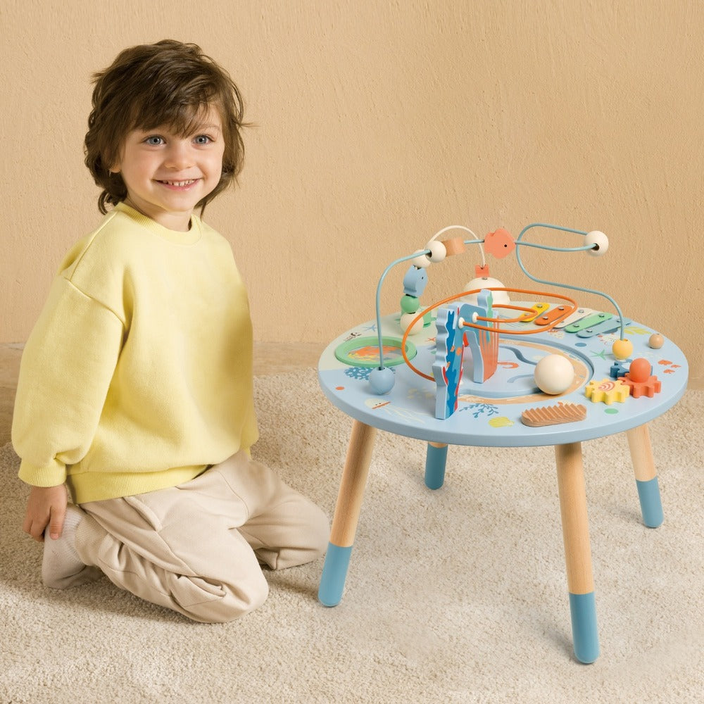 Criança sorridente sentada no chão ao lado da Classic World Mesa de Atividades, que exibe um design com tema oceano e várias funcionalidades interativas.