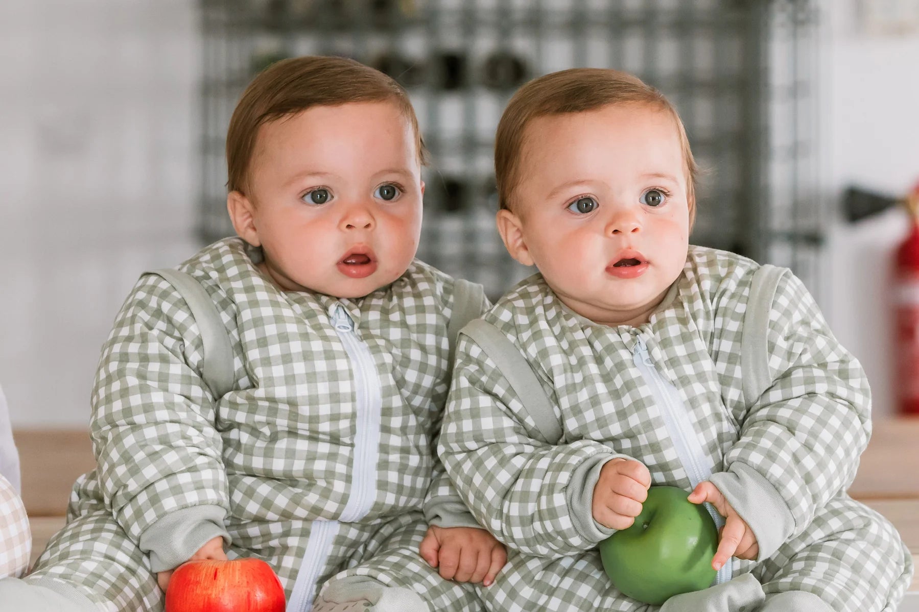Dois bebés gémeos sentados, a usar sacos de dormir MolisandCo TOG 2.5 com mangas e padrão xadrez, segurando frutas.