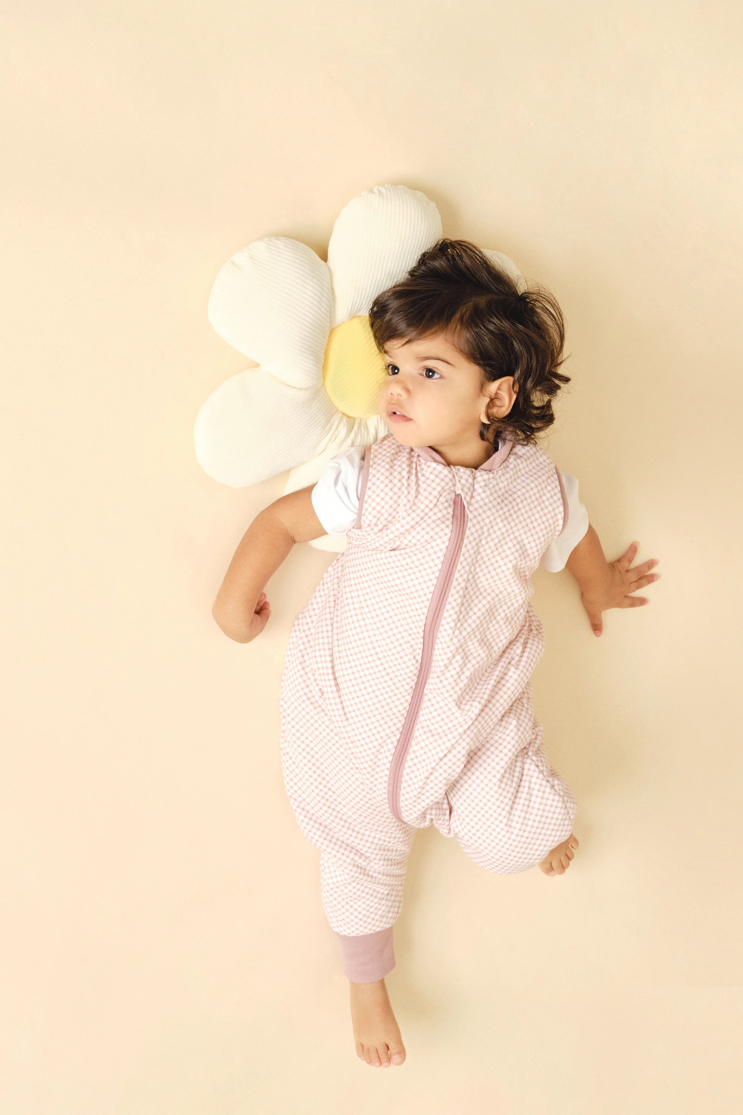 Bebé deitado no chão com o saco de dormir MolisandCo TOG 1.0 VICHY PINK, com as pernas dobradas e a cabeça virada para o lado, ao lado de uma almofada em forma de flor.