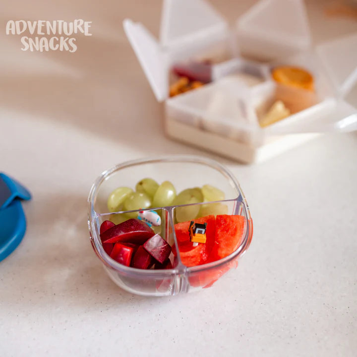 Tigela de lanche Yumbox transparente, em uso, com uvas, maçã e melancia, com o texto "ADVENTURE SNACKS" no canto superior.