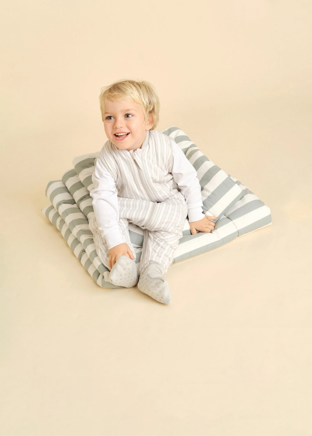 Criança loira sentada sobre almofadas às riscas, a usar o saco de dormir MolisandCo TOG 1.0 Dawn Stripes, com mangas compridas por baixo.