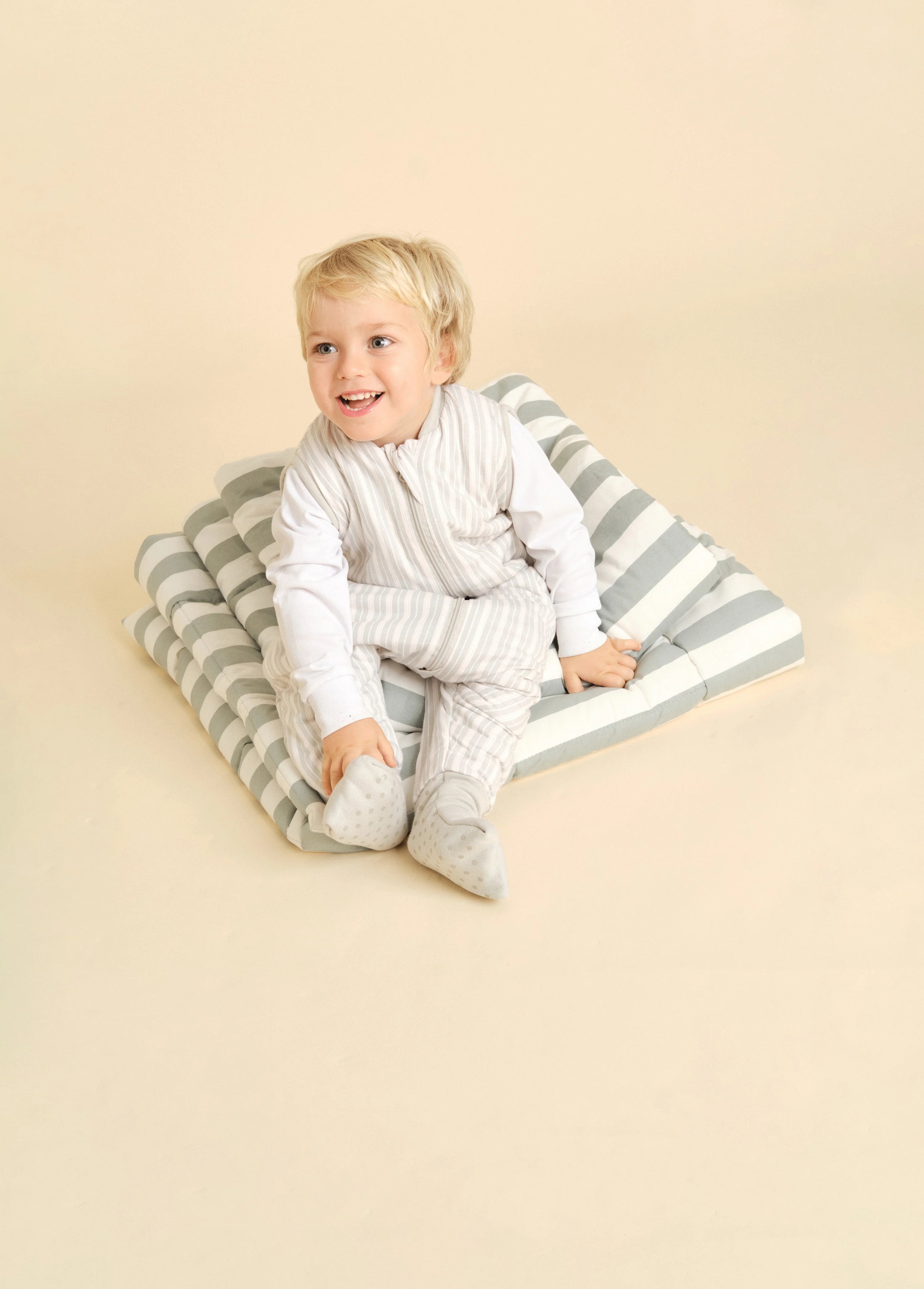 Criança loira sentada sobre almofadas às riscas, a usar o saco de dormir MolisandCo TOG 1.0 Dawn Stripes, com mangas compridas por baixo.