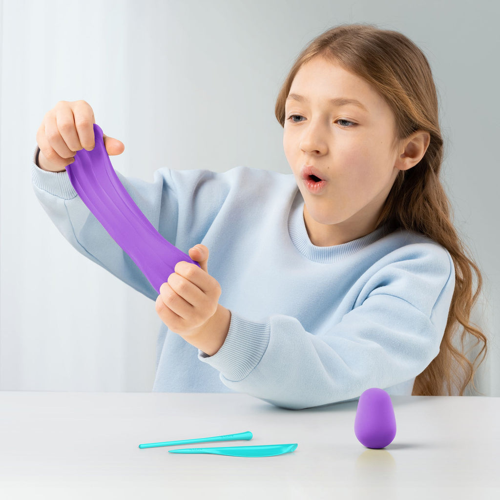 Uma menina a esticar e brincar com a pasta de modelar roxa HEY CLAY, com ferramentas de escultura e uma bola de pasta na mesa.