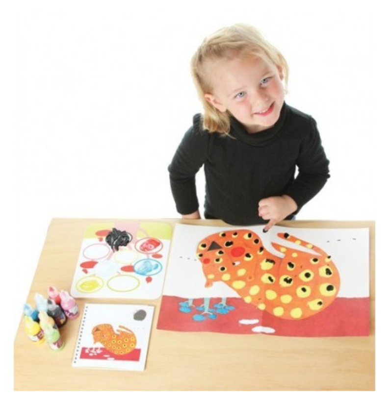 Criança sorridente a pintar com as mãos usando o kit Djeco Atividades c/ linhas, sobre uma mesa de madeira.
