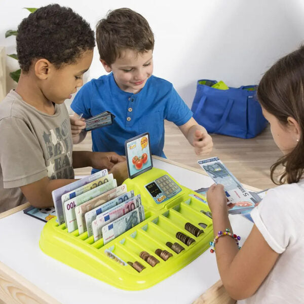 Duas crianças a brincar com o Miniland Euro Shopping, manuseando notas e moedas de euro, com um tablet a exibir um jogo de compras no centro da mesa.