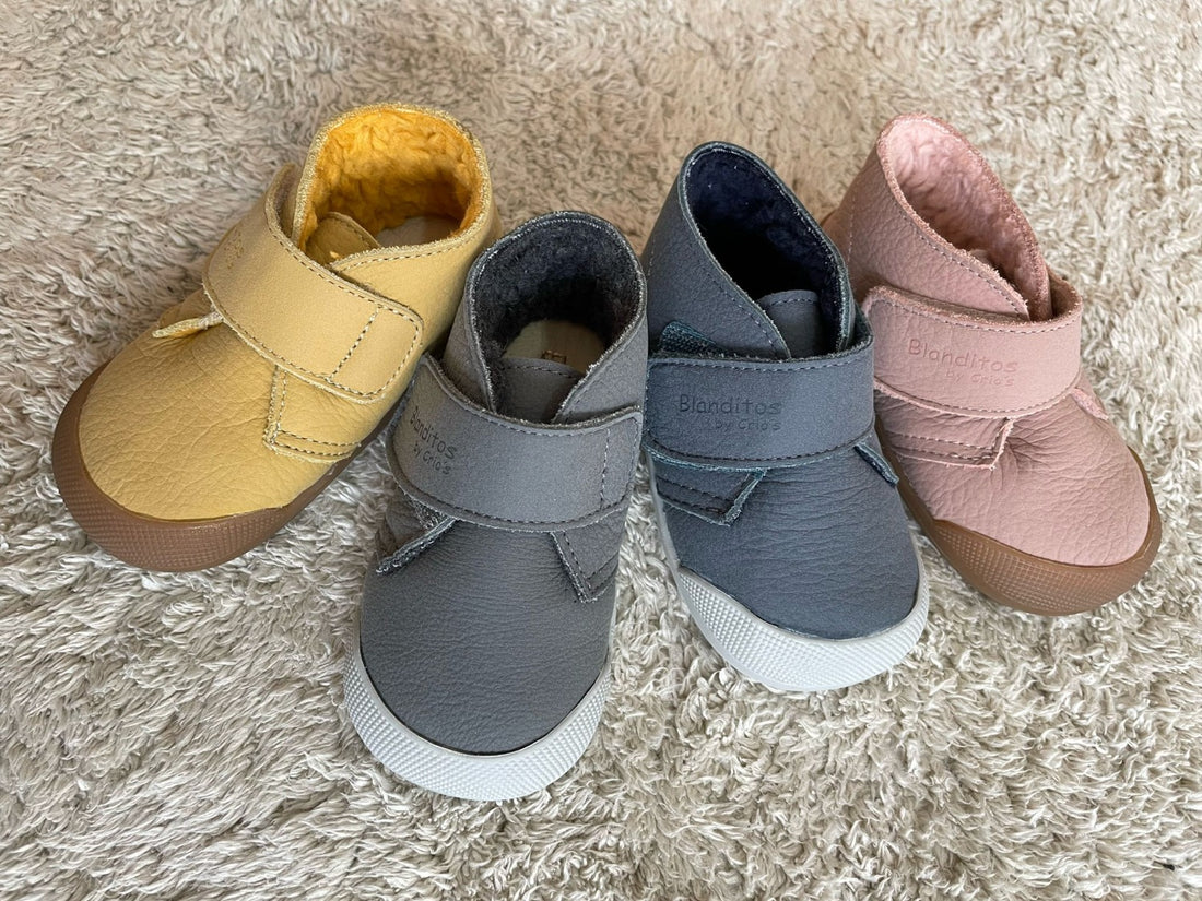 Quatro pares de calçado infantil barefoot para outono/inverno, dispostos sobre uma superfície de neve. Os modelos incluem botinhas de cores azul, vermelho, bege e rosa, com solas flexíveis e detalhes em pelo, perfeitos para manter os pés das crianças quentes e confortáveis.