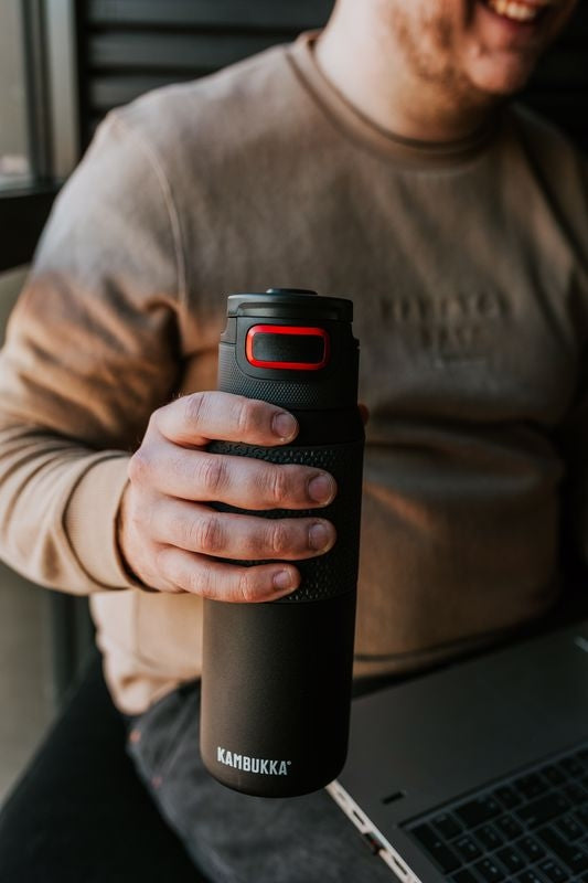 Homem sorridente segura uma garrafa térmica Kambukka ELTON Nightfall preta de 750ml, mostrando a pega texturizada e o detalhe vermelho na tampa, com um portátil ao lado.