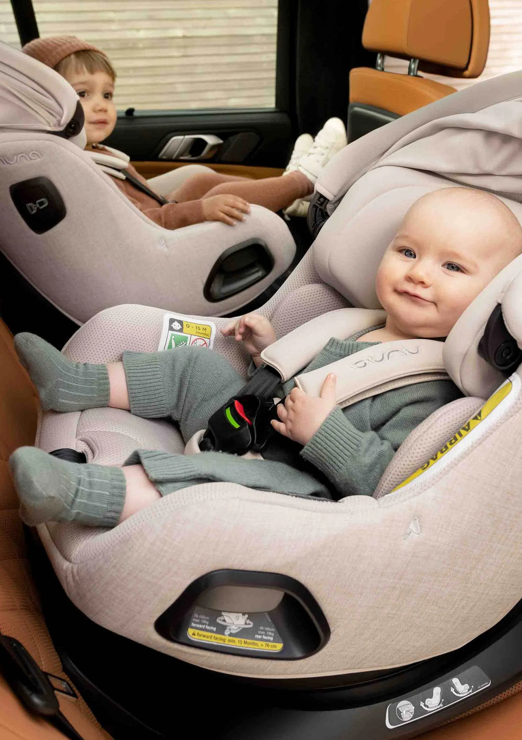 Bebé sorridente sentado na cadeira auto Nuna Pruu Aire, vestido de verde, com um irmão mais velho visível ao fundo no banco do carro.