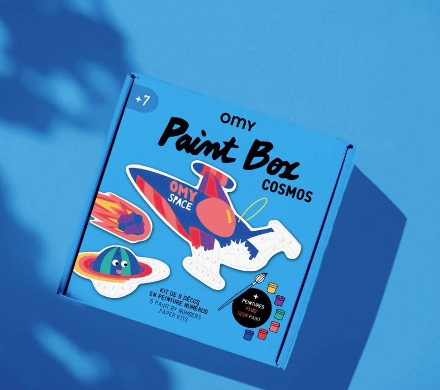 Caixa OMY Paint Box Cosmos para colorir, com ilustrações de foguetão, planeta e indicação de idade recomendada (+7 anos).