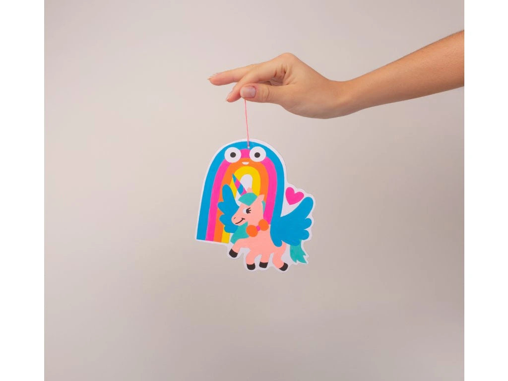 Decoração de unicórnio e arco-íris pintada, suspensa por uma mão, mostrando o resultado final do kit OMY Lily.