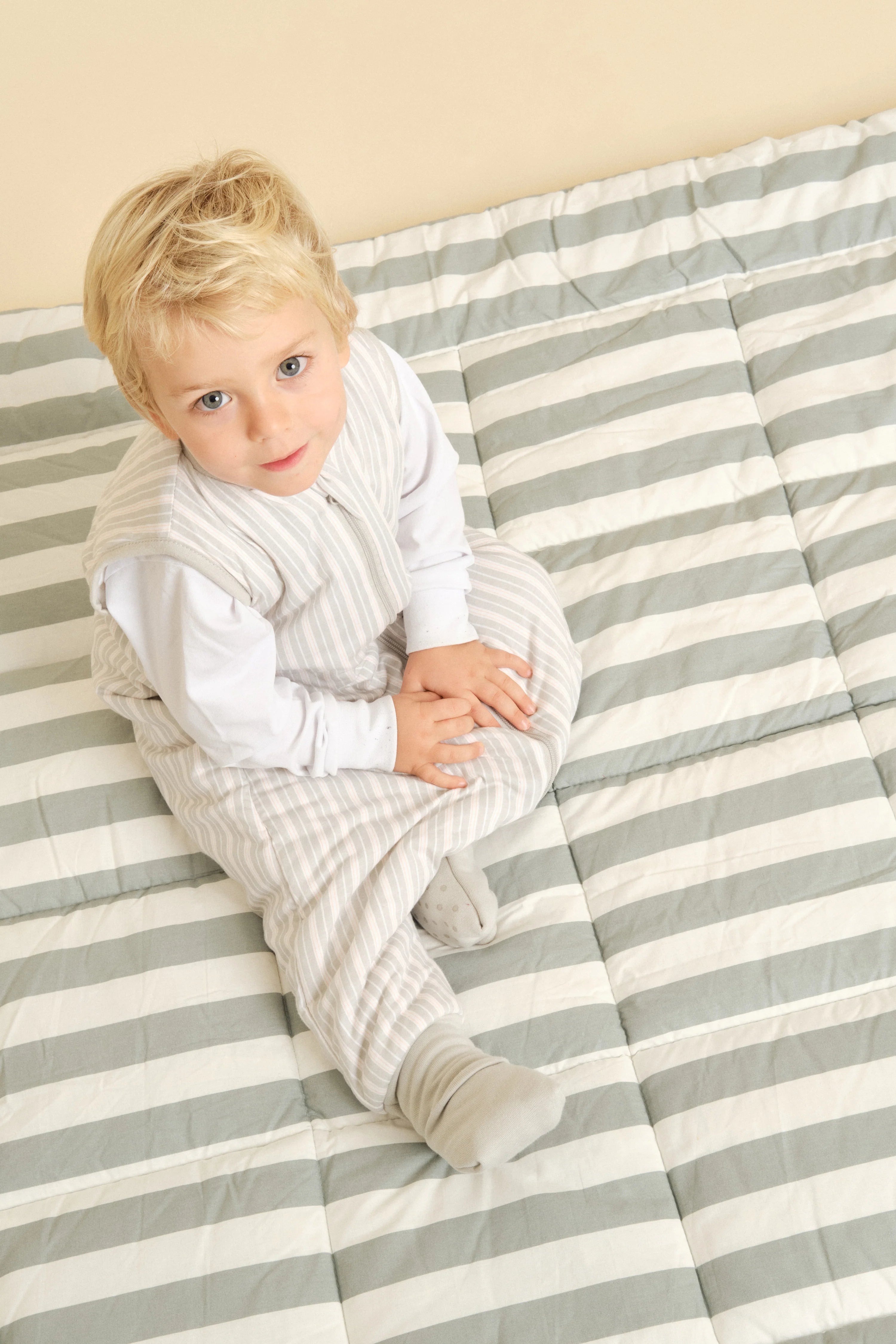 Criança loira deitada numa cama às riscas, a usar o saco de dormir MolisandCo TOG 1.0 Dawn Stripes, olhando para cima.